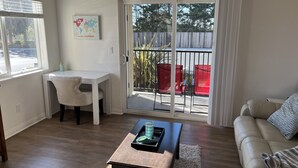 A living room with a white sofa, a wooden coffee table, a white desk with a chair, and a sliding glass door leading to an outdoor patio with red chairs. - Luxury Pismo Beach condo close to shopping and mins drive to the beach. (Pismo Beach)