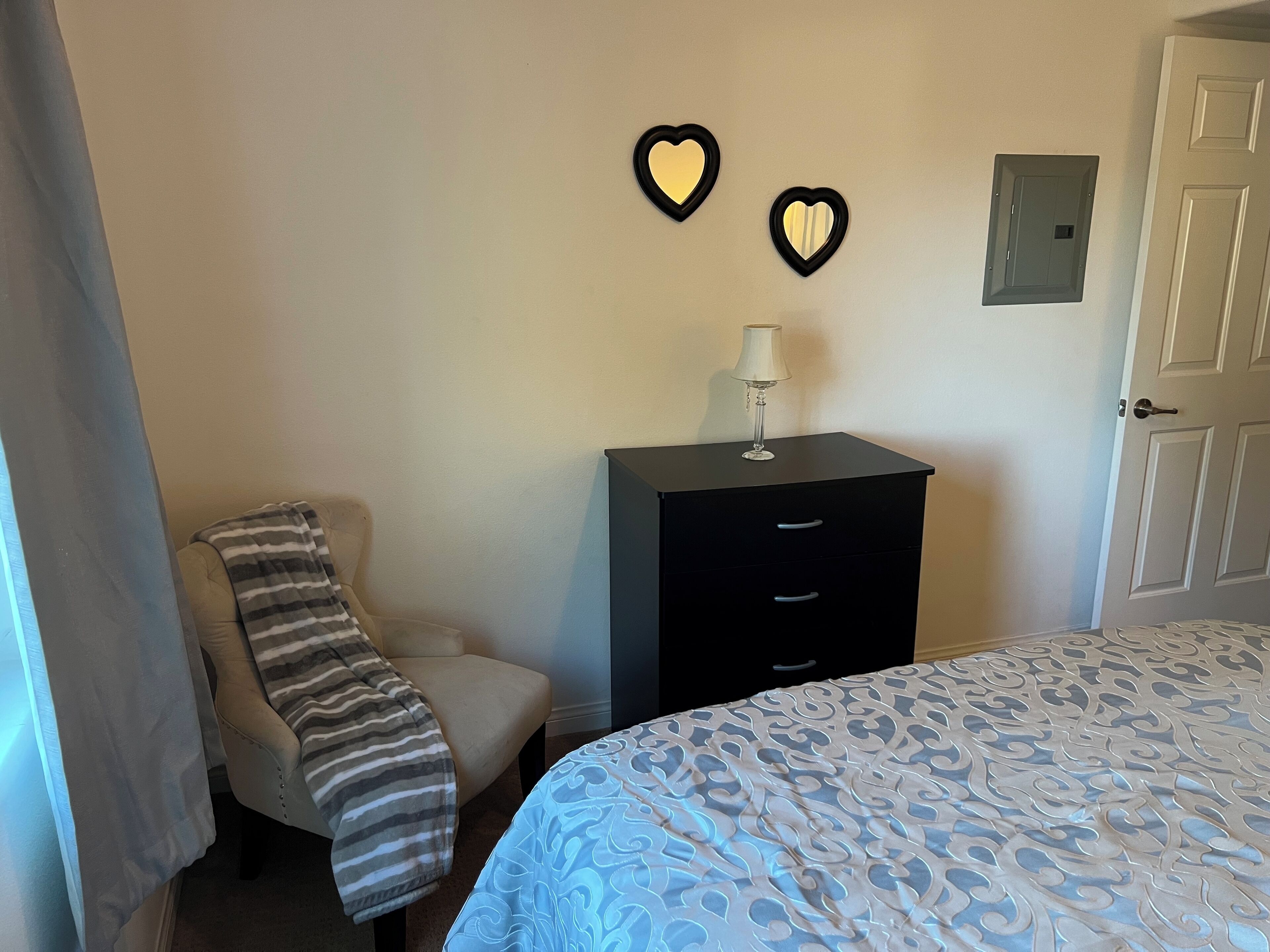 Una camera da letto con un letto, una sedia con coperta a righe, una cassettiera con una lampada e due specchi a forma di cuore sulla parete.
