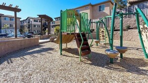 A playground with a slide and climbing structures, surrounded by a gravel area and residential buildings in the background. - Luxury Pismo Beach condo close to shopping and mins drive to the beach. (Pismo Beach)