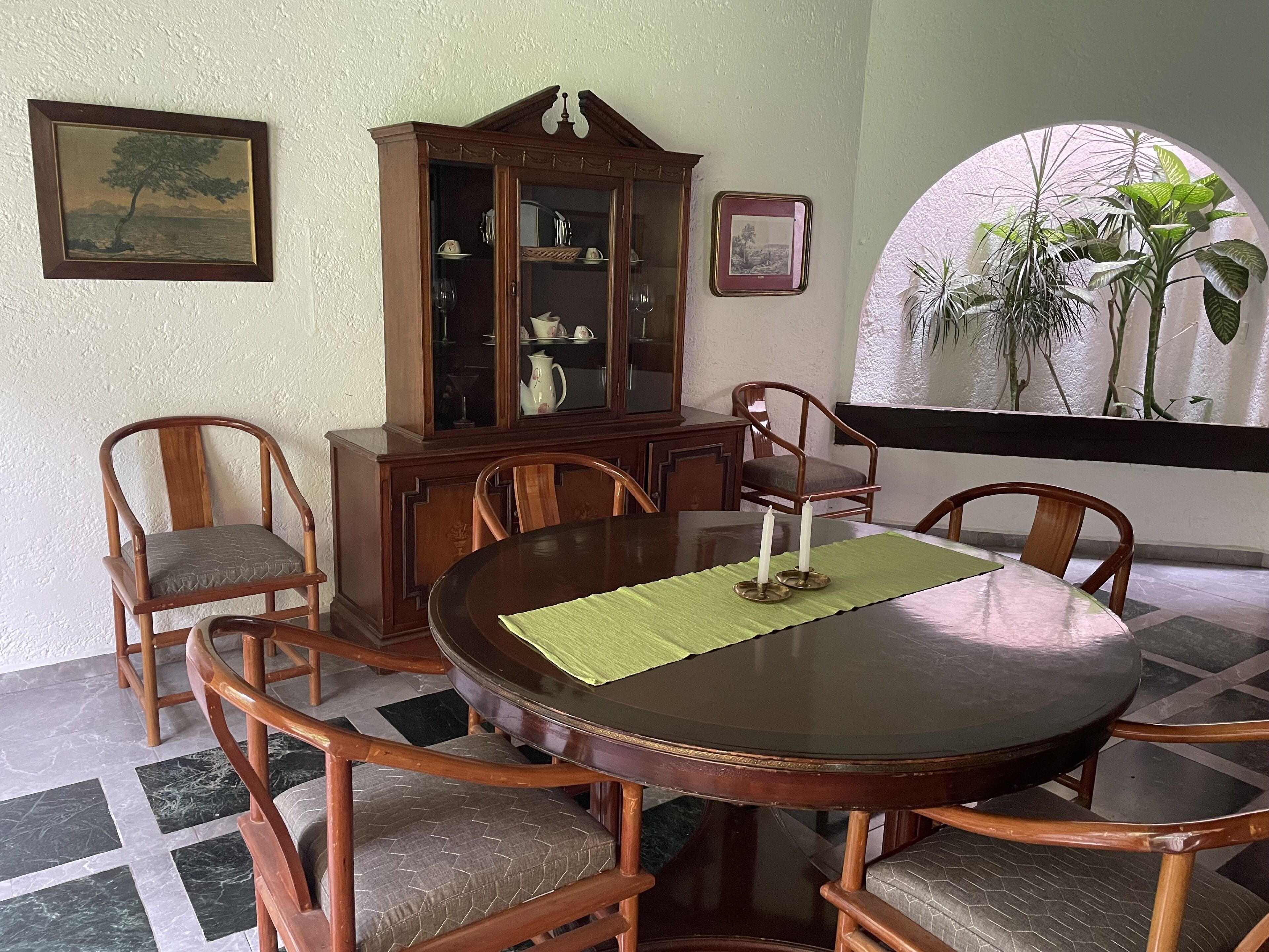 A dining area with a round table, wooden chairs, a cabinet, and a window with plants.