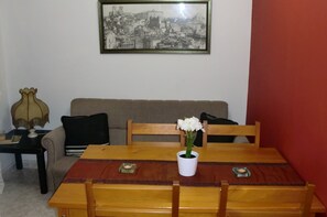 A dining area with a wooden table and chairs, a framed cityscape picture on the wall, a sofa, and a vase with flowers.