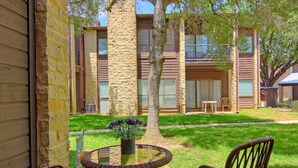 A patio with a round table and chairs, a potted plant, and a view of a building with balconies and windows. - Go with the Flow! CW B103 by RedAwning (New Braunfels)