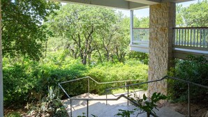 A covered outdoor area with stone pillars, a metal railing, and a view of a lush garden with trees and shrubs. - Parker River Haus Vg106 3 Bedroom Condo by RedAwning (New Braunfels)