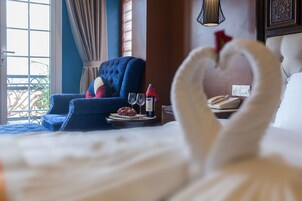A hotel room with a bed, a blue armchair, a dining table with a plate of grapes and a bottle of wine, and a telephone on the nightstand.