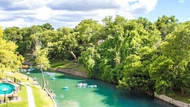 Ein Fluss mit Booten, umgeben von dichtem Baumbestand und einem Schwimmbereich mit Pool und Liegestühlen.