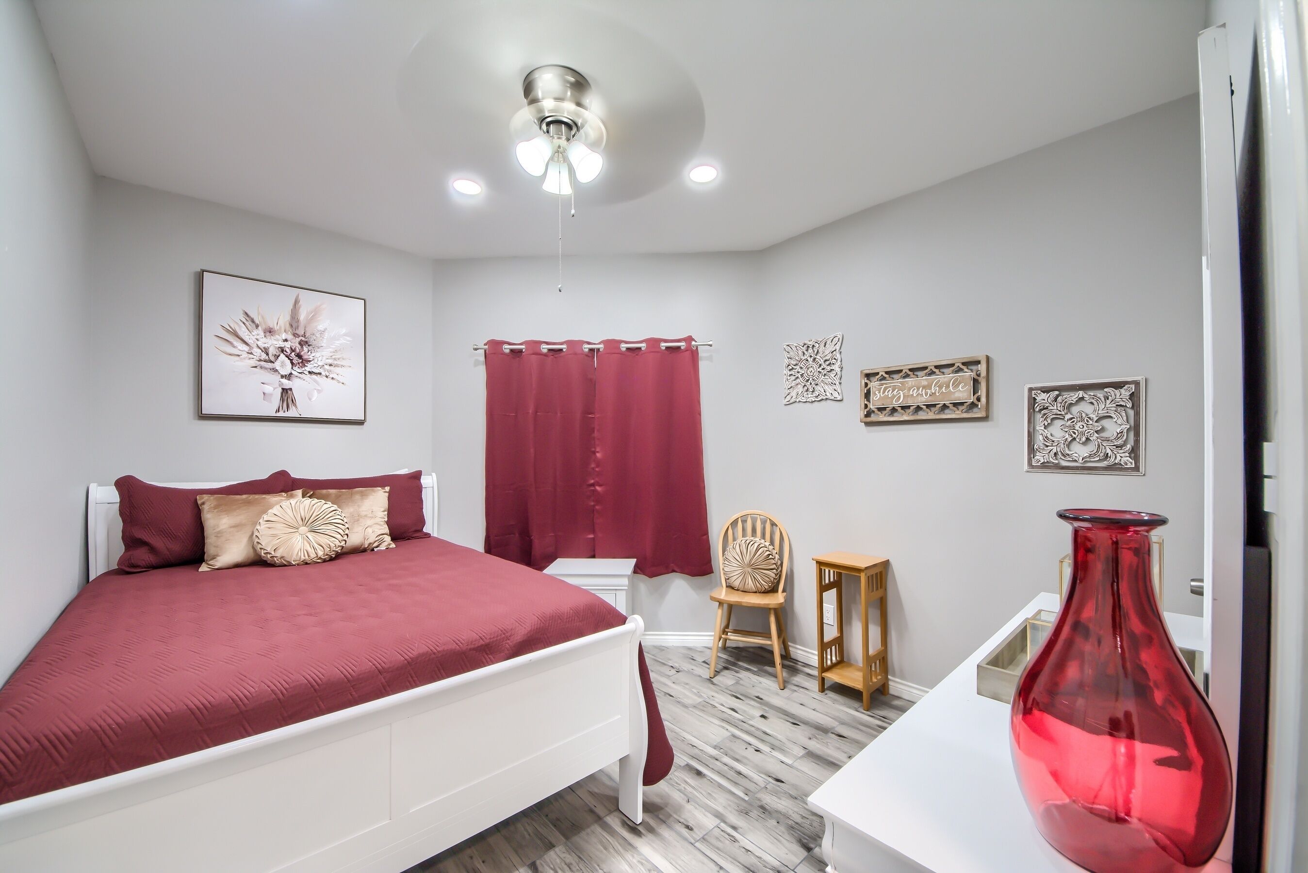 Un dormitorio con cama, una silla, una mesita, un jarrón rojo y obras de arte en las paredes.