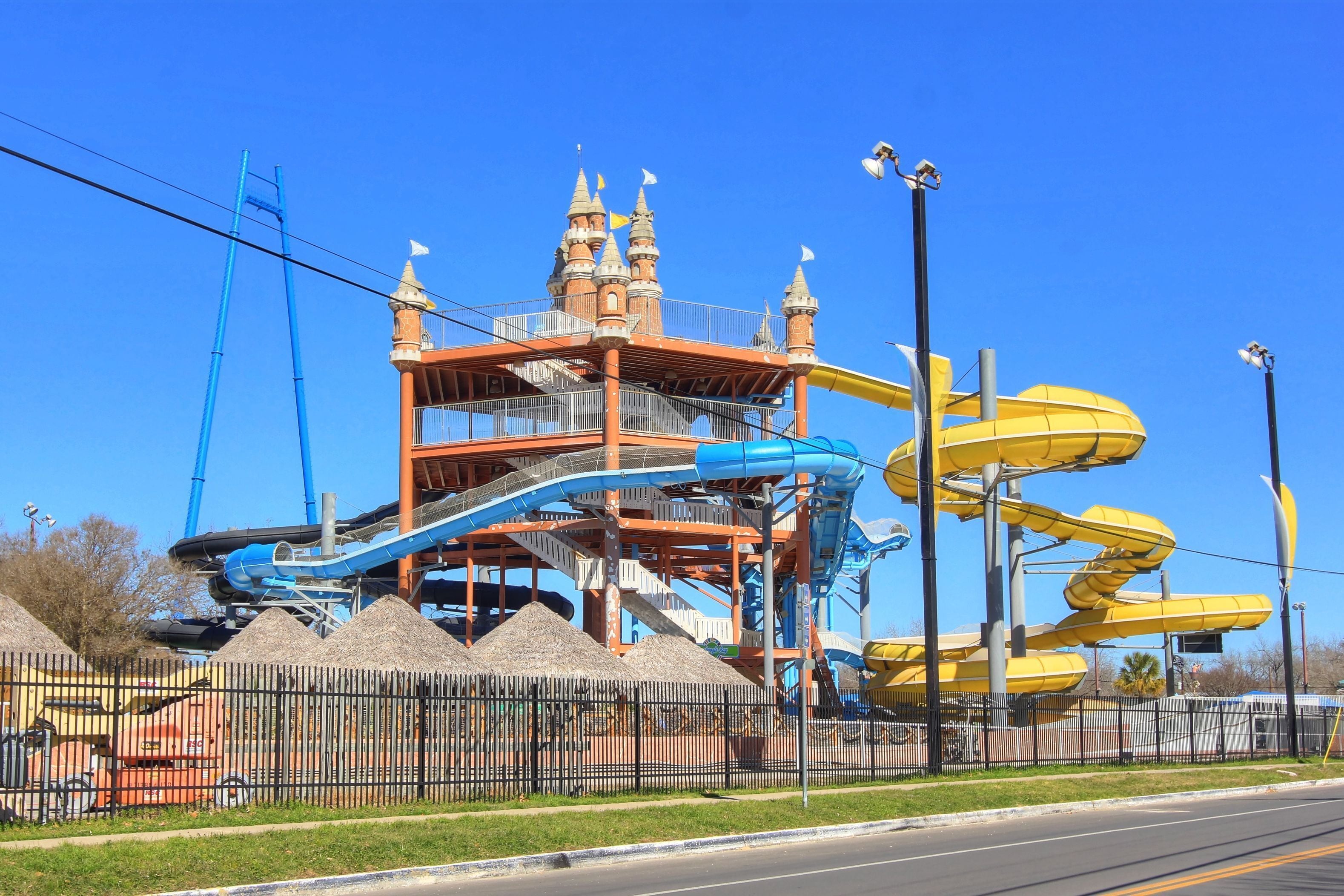 A water park with a castle-like structure, multiple water slides, and a fenced area with a trash bin.