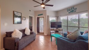 A living room with a grey sofa, a teal coffee table, and a blue armchair. There is a ceiling fan, a TV, and a window with blinds. - Inverness Condos #302 by RedAwning (New Braunfels)