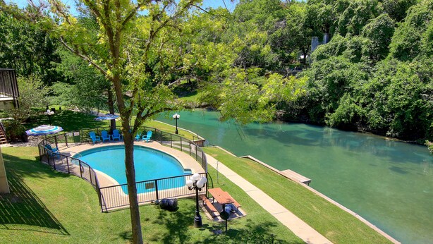 A swimming pool surrounded by a deck with chairs and a table, a grassy area, and a river. - Inverness Condos #302 by RedAwning (New Braunfels)