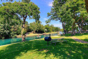 Enjoy a serene riverside retreat with a barbecue grill and picnic tables amidst lush greenery and mature trees.