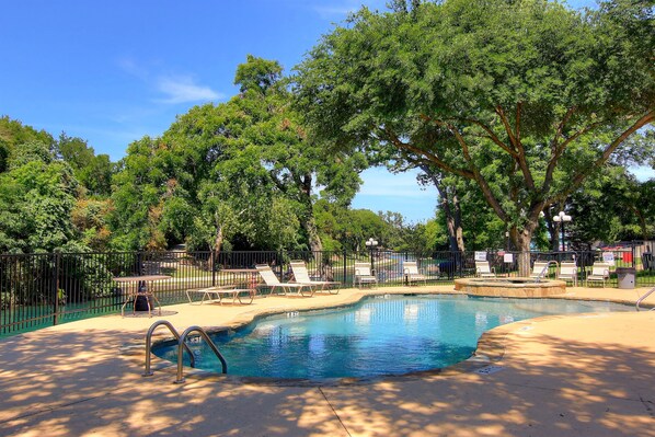 Relax by the serene pool surrounded by lush greenery and mature trees, with lounge chairs and a dining area.