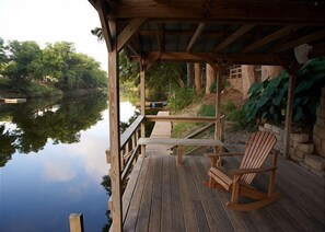 A wooden deck with a table and chairs overlooking a calm body of water, surrounded by greenery. - Leaning Oak PRD923 by RedAwning (New Braunfels)