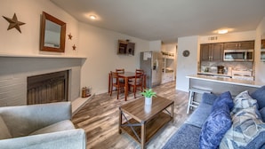 A modern living room with a fireplace, a dining area with wooden chairs, and a kitchen with a microwave. - Grandma's River Haus       Rrc A108 1 Bedroom Condo by RedAwning (New Braunfels)