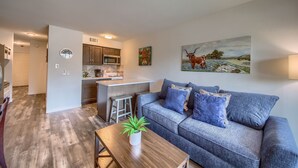 A modern living room with a grey sofa, a wooden coffee table with a plant, and a kitchen in the background. - Grandma's River Haus       Rrc A108 1 Bedroom Condo by RedAwning (New Braunfels)