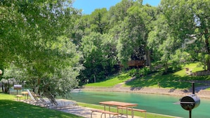 A picnic area with a table and benches by a calm river, surrounded by lush greenery and a wooden deck.