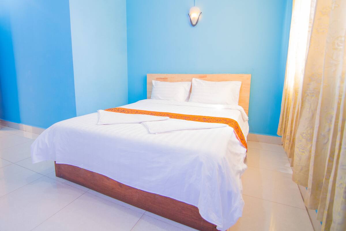 A neatly made bed with white linens and an orange decorative throw, a wooden headboard, and a single bedside lamp.