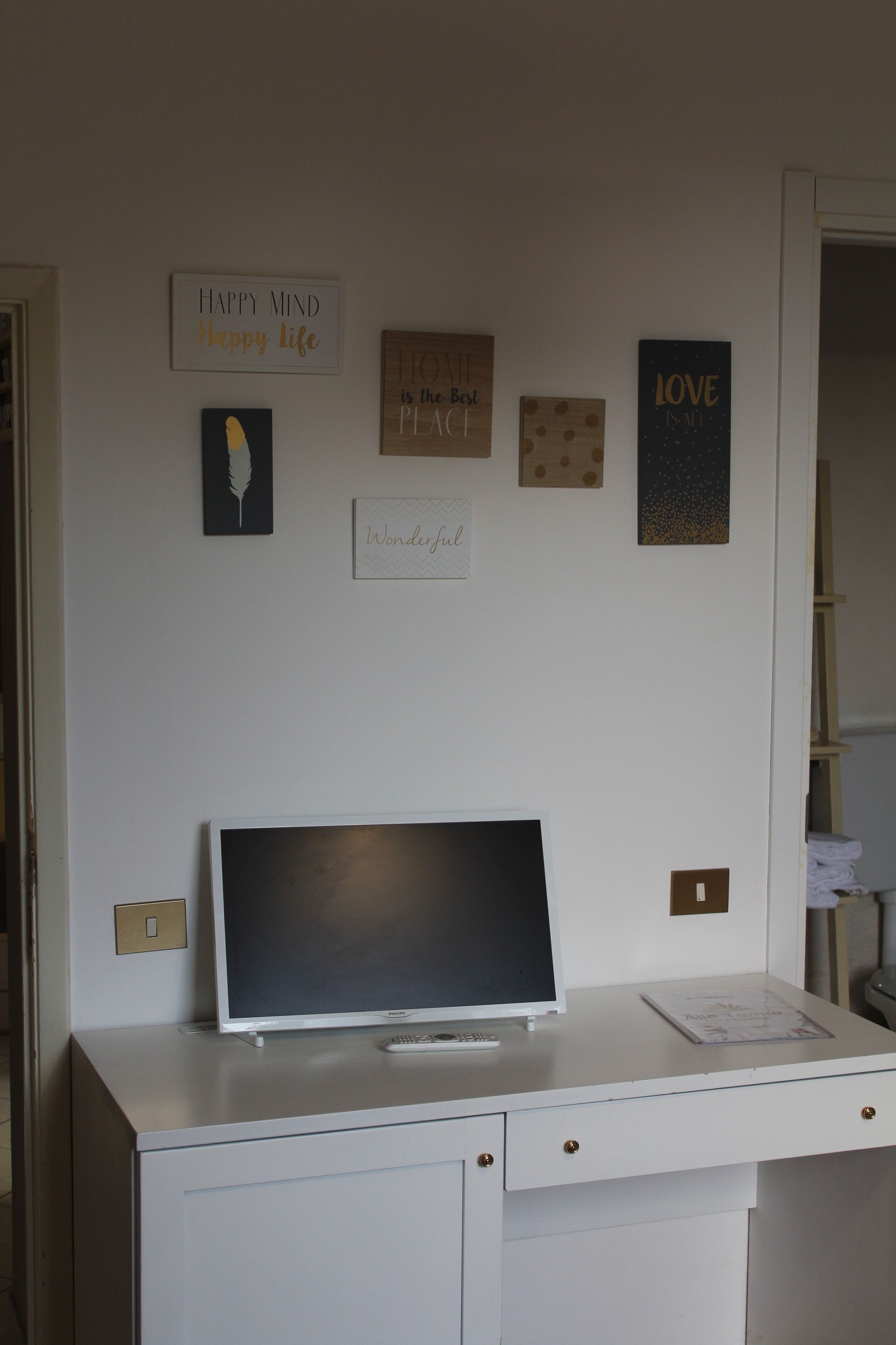 Una scrivania con un monitor del computer, un telecomando e un libro. Decorazioni a parete tra cui una piuma, un cuore e un cartello con la scritta "HAPPY MIND Happy life".