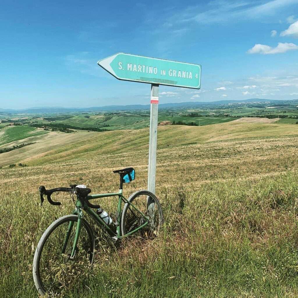 A signpost with 'S. MARTINO IN GRANIA' pointing to the right, a bicycle leaning against it, and a grassy landscape with hills in the background.