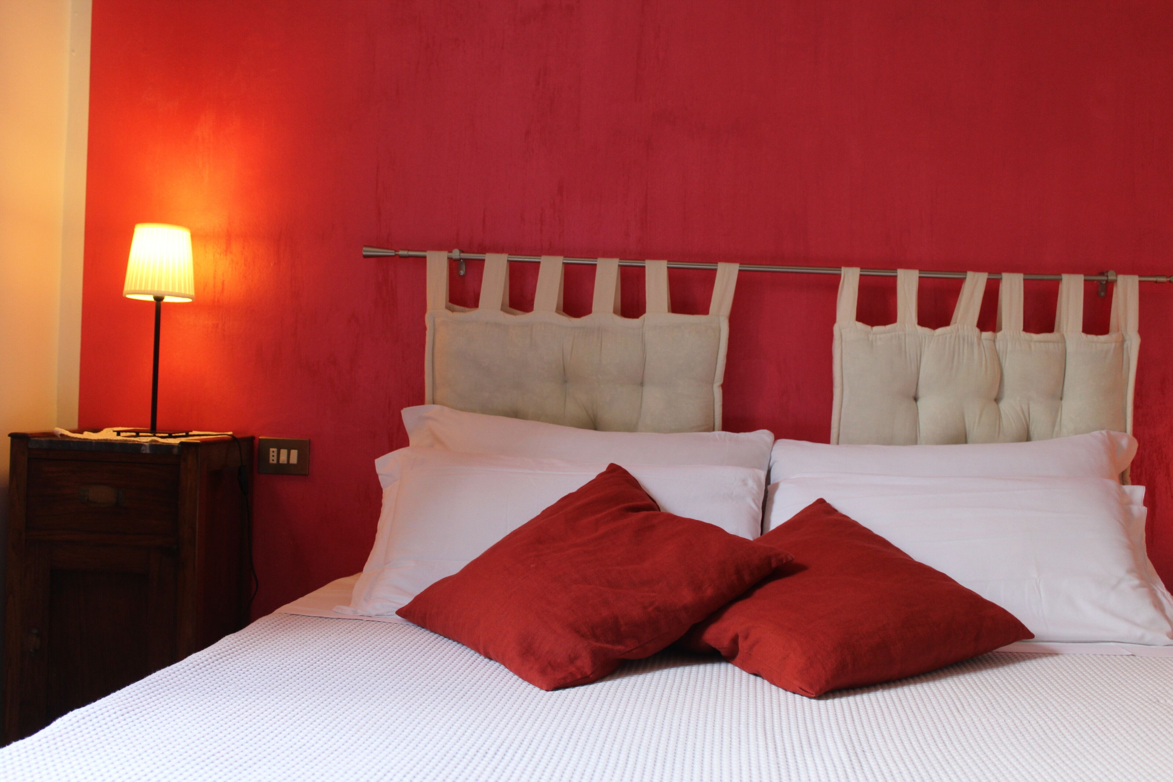 Una cama con ropa blanca y dos almohadas rojas, una lámpara de noche y una mesita de madera.