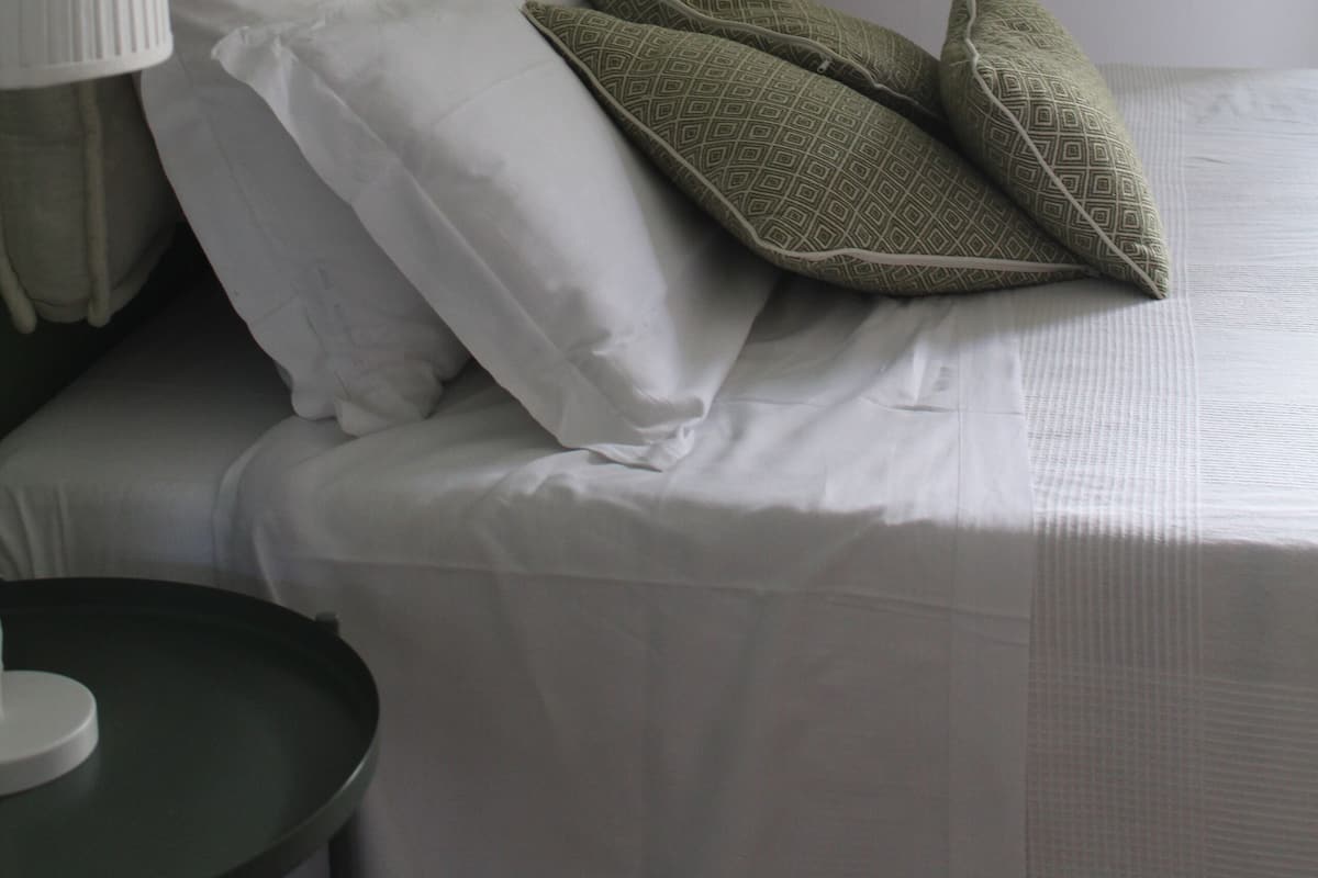 Een keurig opgemaakt bed met witte lakens en kussens met een groen patroon, een bedlamp en een klein rond tafeltje.