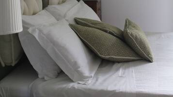 Uma cama bem arrumada com lençóis brancos e almofadas estampadas em verde, uma luminária de cabeceira e uma pequena mesa redonda.