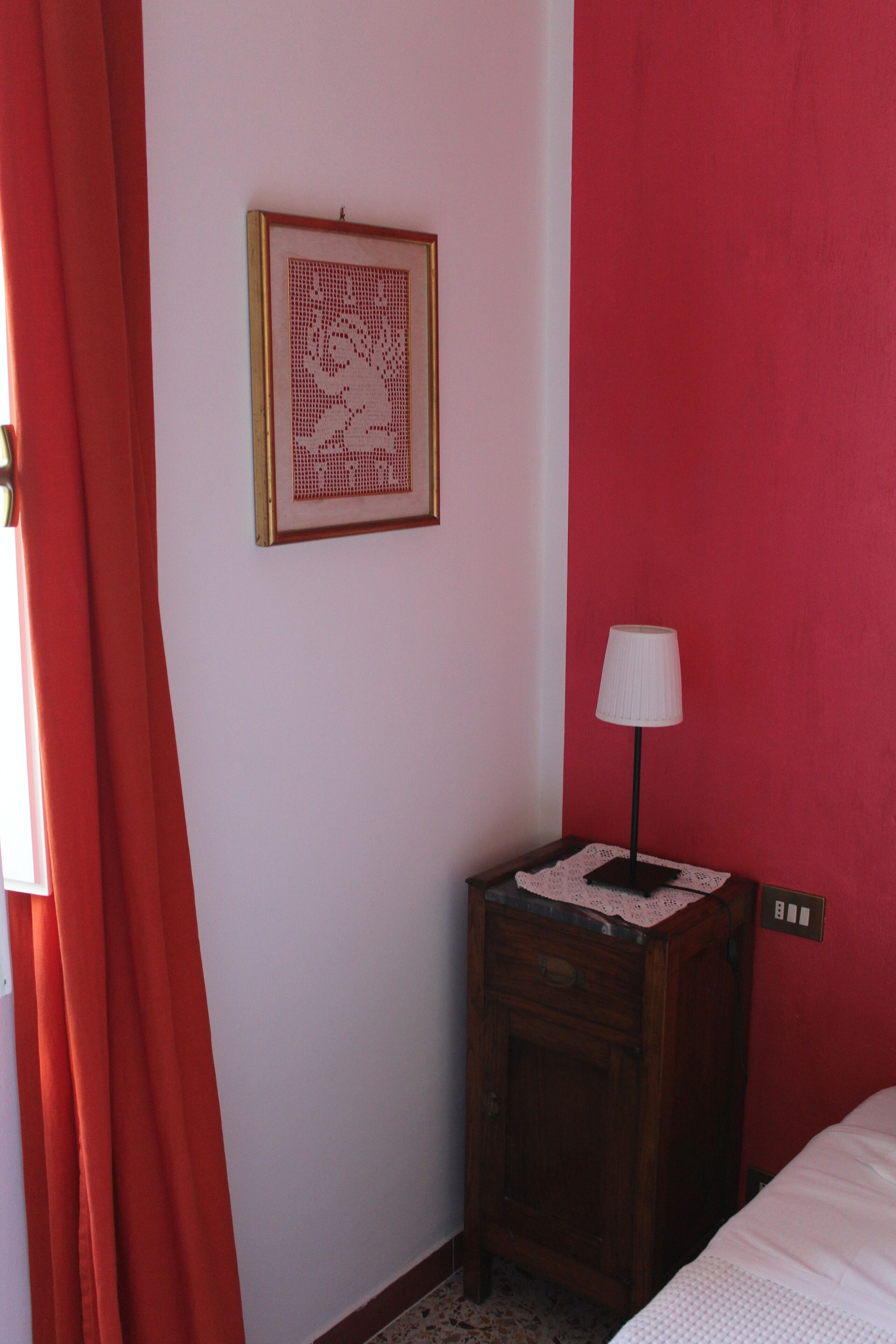 Ein Zimmer mit einem Bett, einem Nachttisch mit Lampe, einem gerahmten Kunstwerk und einer roten Wand.