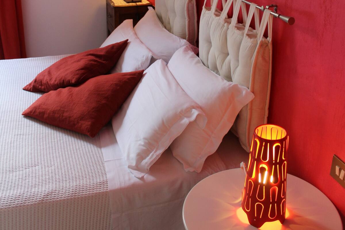 Een net opgemaakt bed met witte en bruine kussens, een bedlampje en een rode muur.