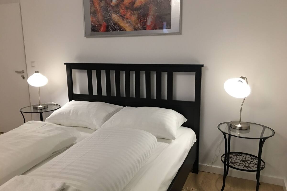 A neatly made bed with white linens, two bedside lamps, a framed abstract painting above the bed, and a patterned rug on the floor.