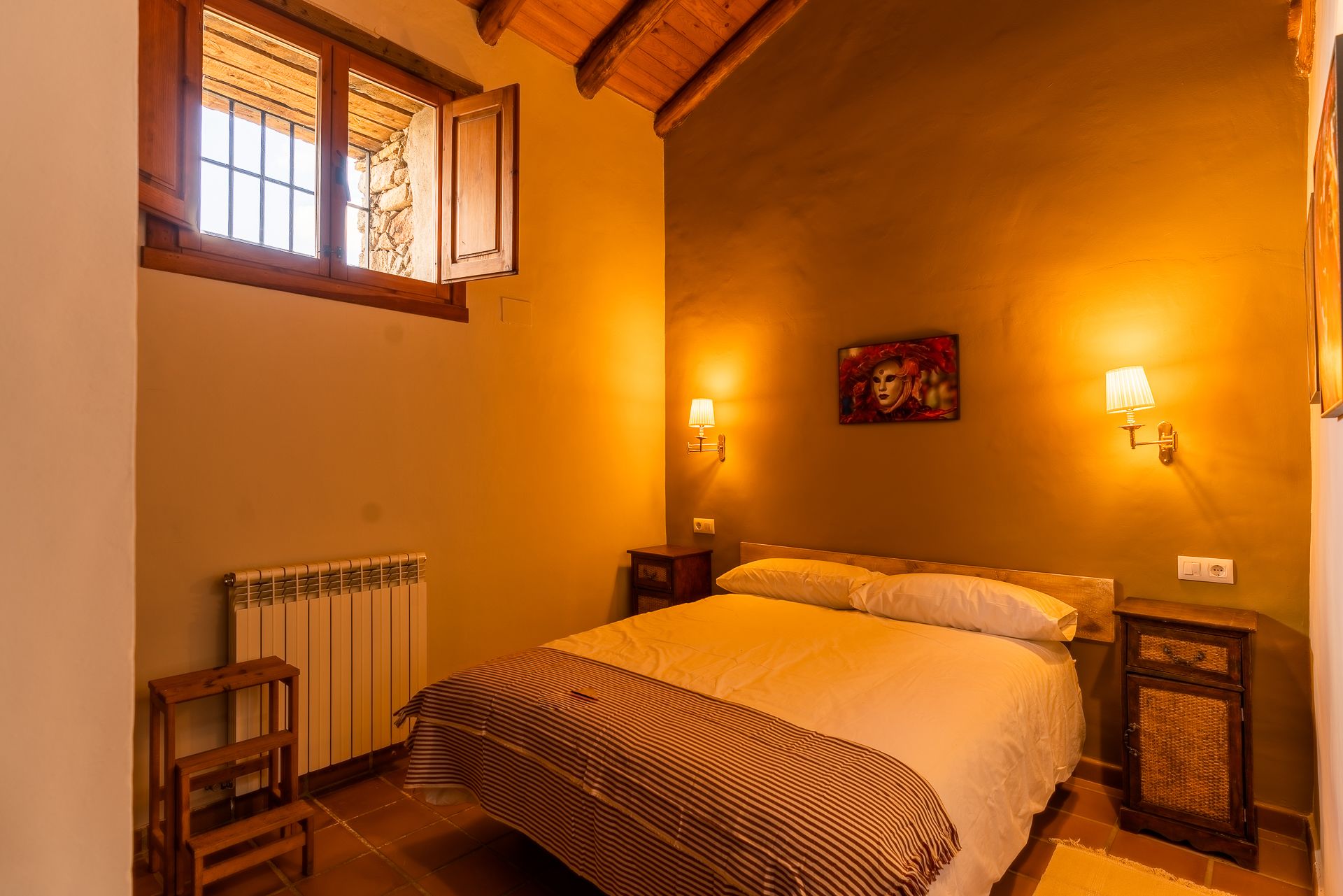 A cozy bedroom with a bed, two bedside tables, a radiator, and a window with wooden shutters.
