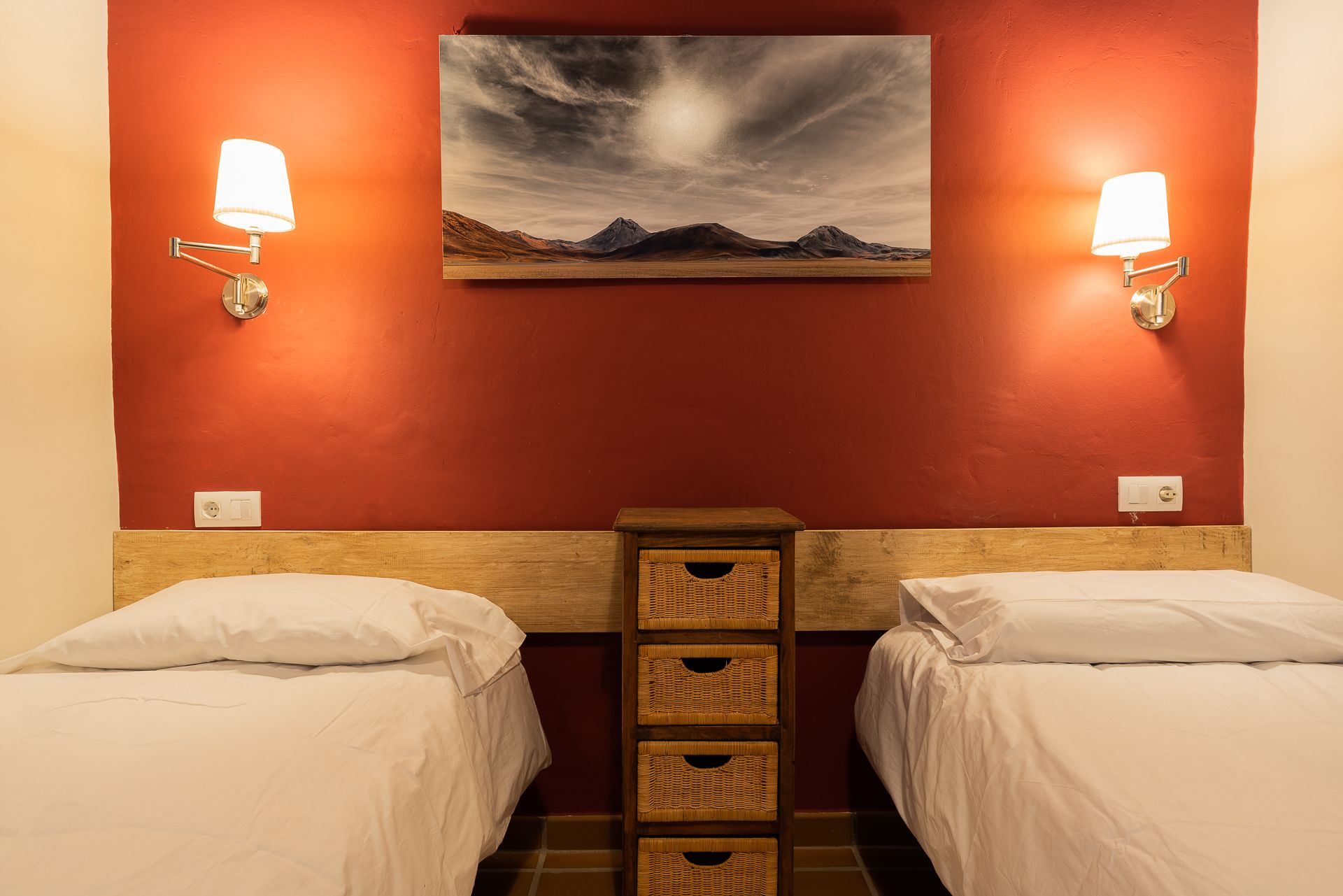 A hotel room with two beds, a wooden headboard, a wall-mounted painting of a landscape, and wall-mounted reading lights.