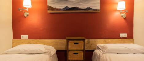 A hotel room with two beds, a wooden headboard, a wall-mounted painting of a landscape, and wall-mounted reading lights.