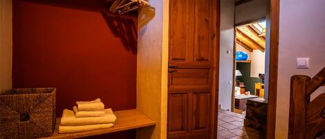 A bathroom with a wooden door, a bench with towels, and a shelf with hanging items.