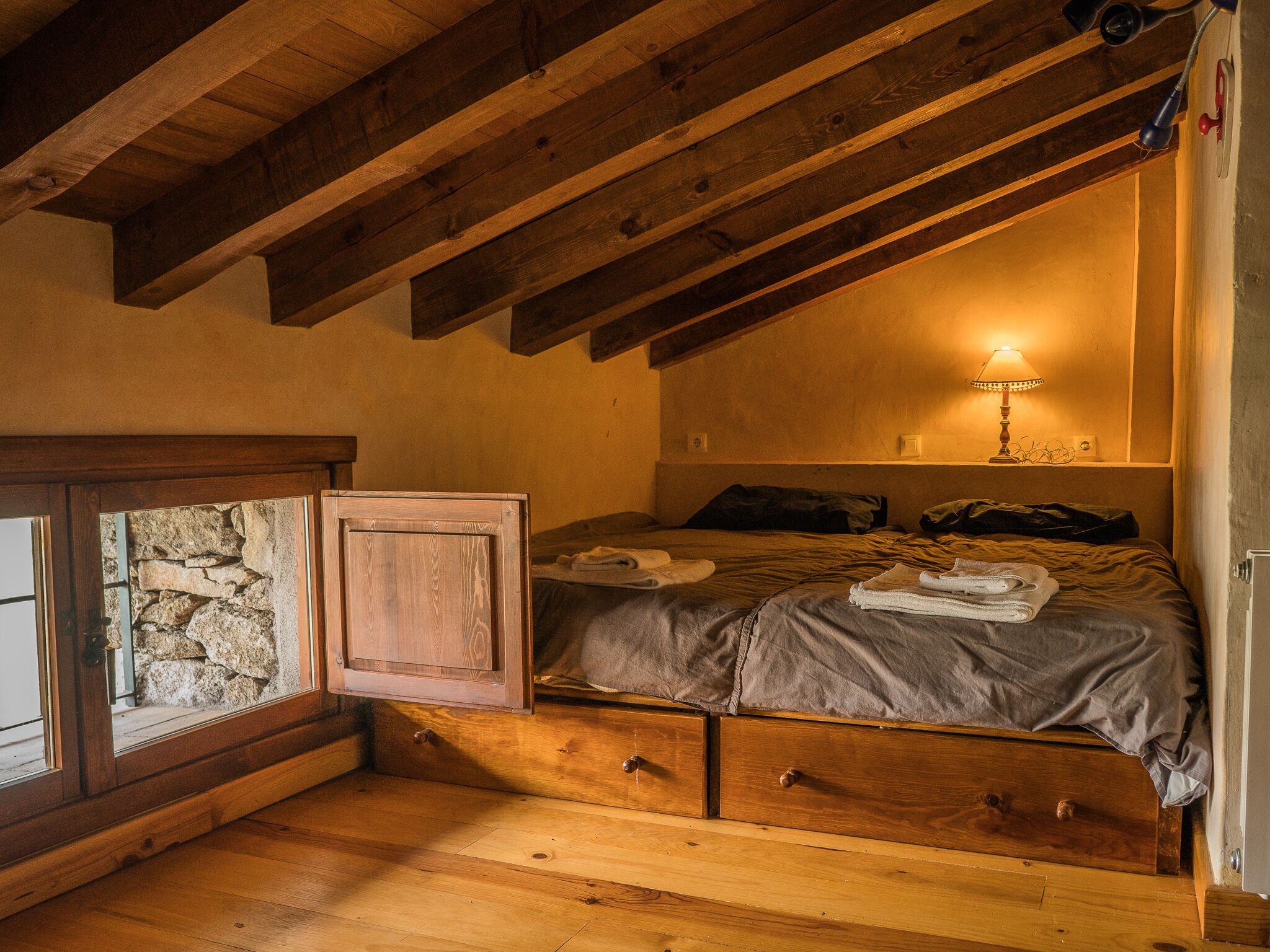 A cozy bedroom with a wooden ceiling, a bed with a grey blanket, a bedside lamp, and a stone wall.