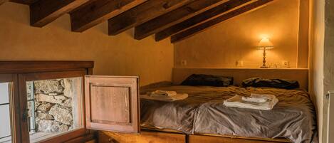A cozy bedroom with a wooden ceiling, a bed with a grey blanket, a bedside lamp, and a stone wall.