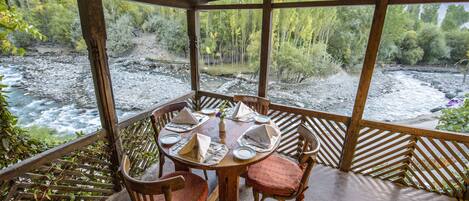 Une table en bois, dressée pour quatre personnes, avec des chaises, donnant sur une rivière et une zone boisée.