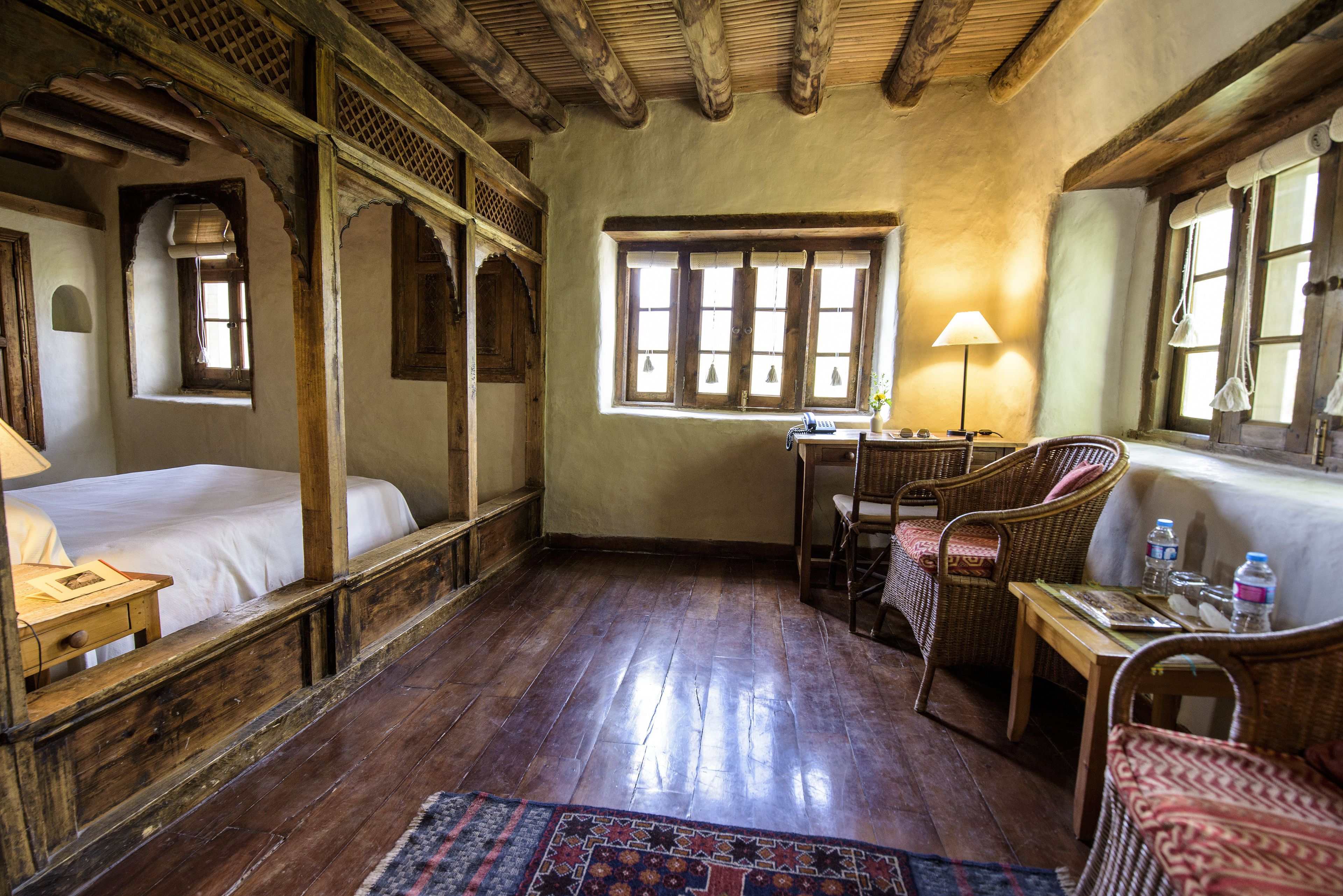 Una habitación tradicional con suelo de madera, una cama, un escritorio con lámpara y muebles de mimbre.