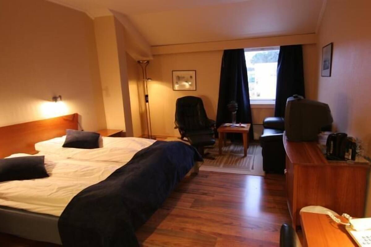 A hotel room with a bed, a desk with a telephone, a chair, a television, and a window with curtains.