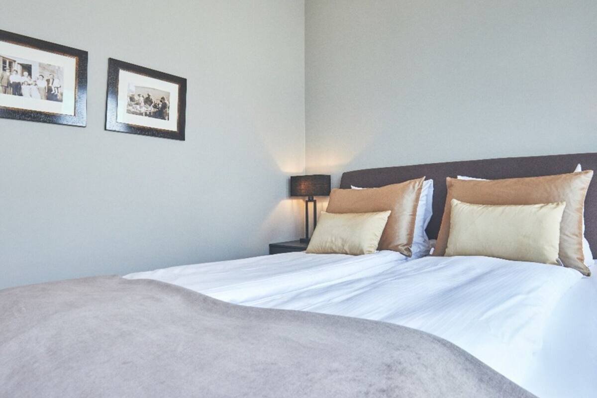 A neatly made bed with a large beige blanket, two bedside tables with lamps, and two framed photographs on the wall.