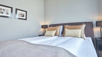 A neatly made bed with a large beige blanket, two bedside tables with lamps, and two framed photographs on the wall.