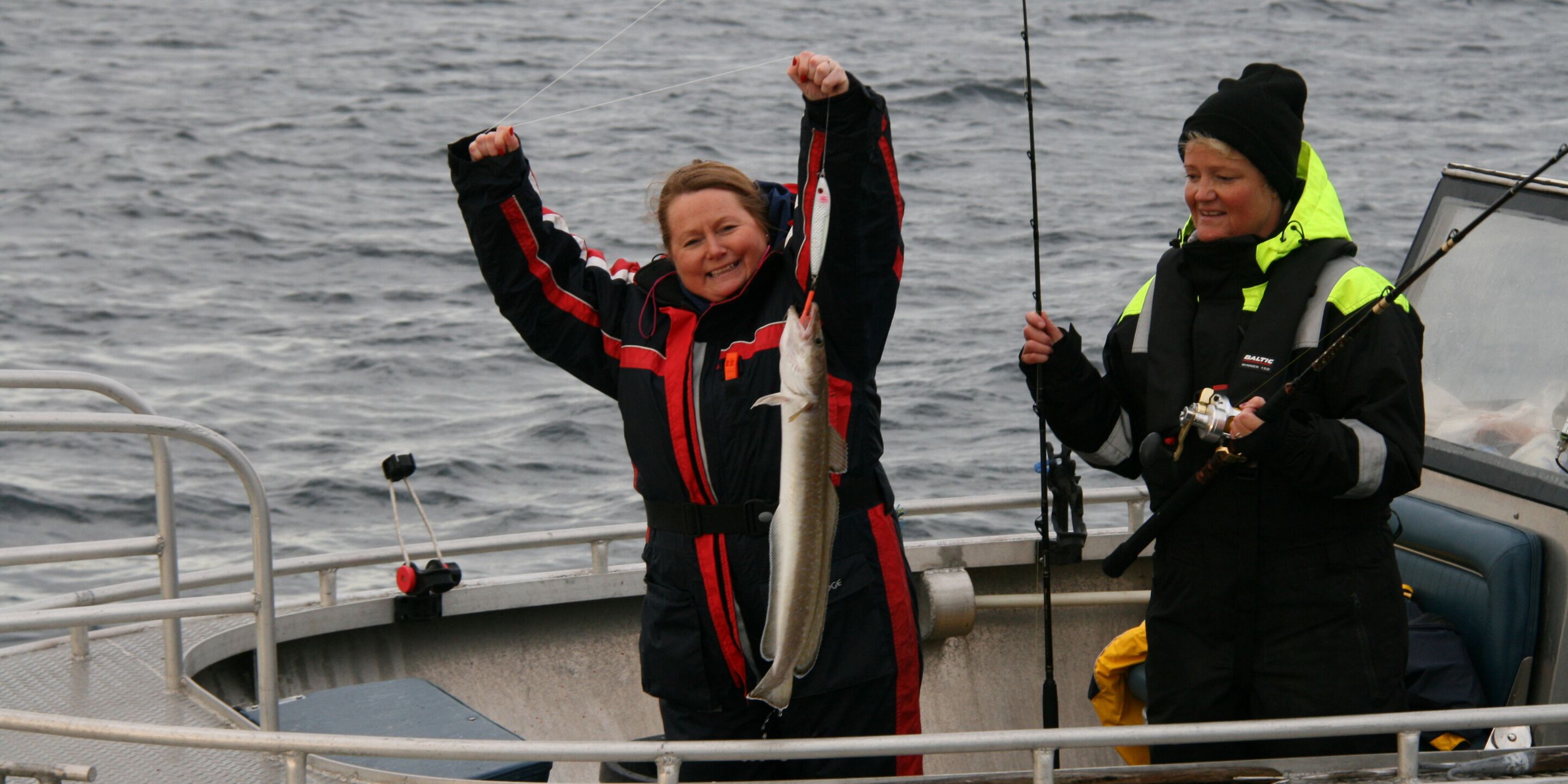 Two people on a boat holding a fish, with a railing and water in the background.