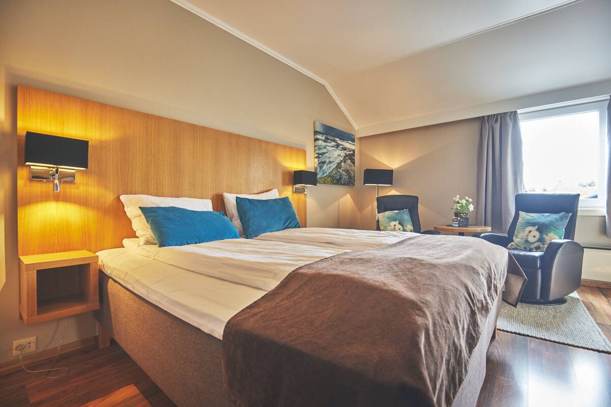 A modern hotel room with a large bed, wooden headboard, and a view of the outdoors.