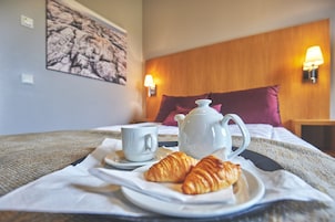 A neatly made bed with a white duvet, a white teapot, a white cup, and a croissant on a white plate, all set on a white tablecloth.