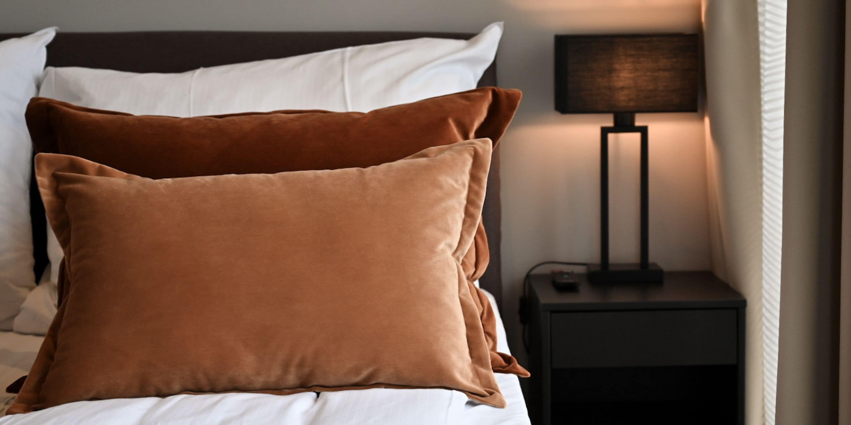 Ein ordentlich bezogenes Bett mit braunen Kissen und einer weißen Decke. Neben dem Bett steht ein schwarzer Nachttisch mit einer Lampe.