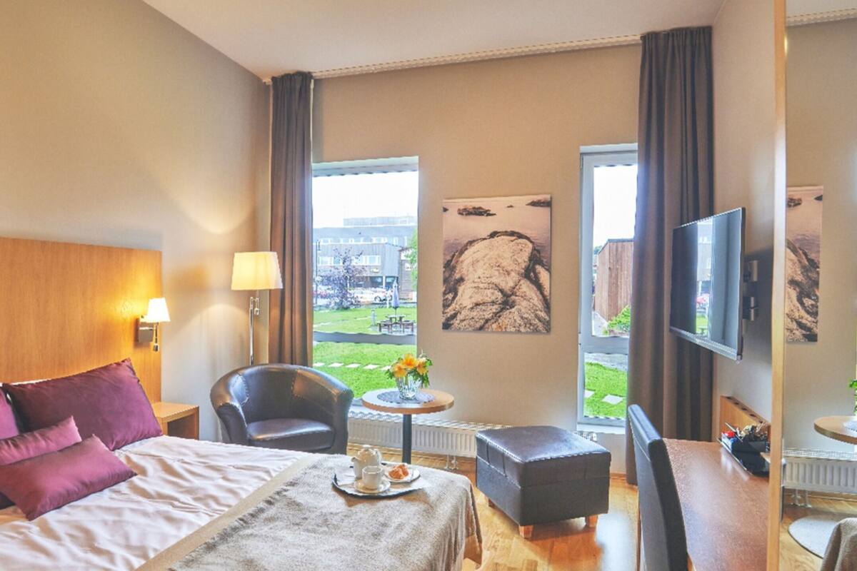 A hotel room with a bed, a TV, a dining area with a table and chairs, and a view of the outdoors.