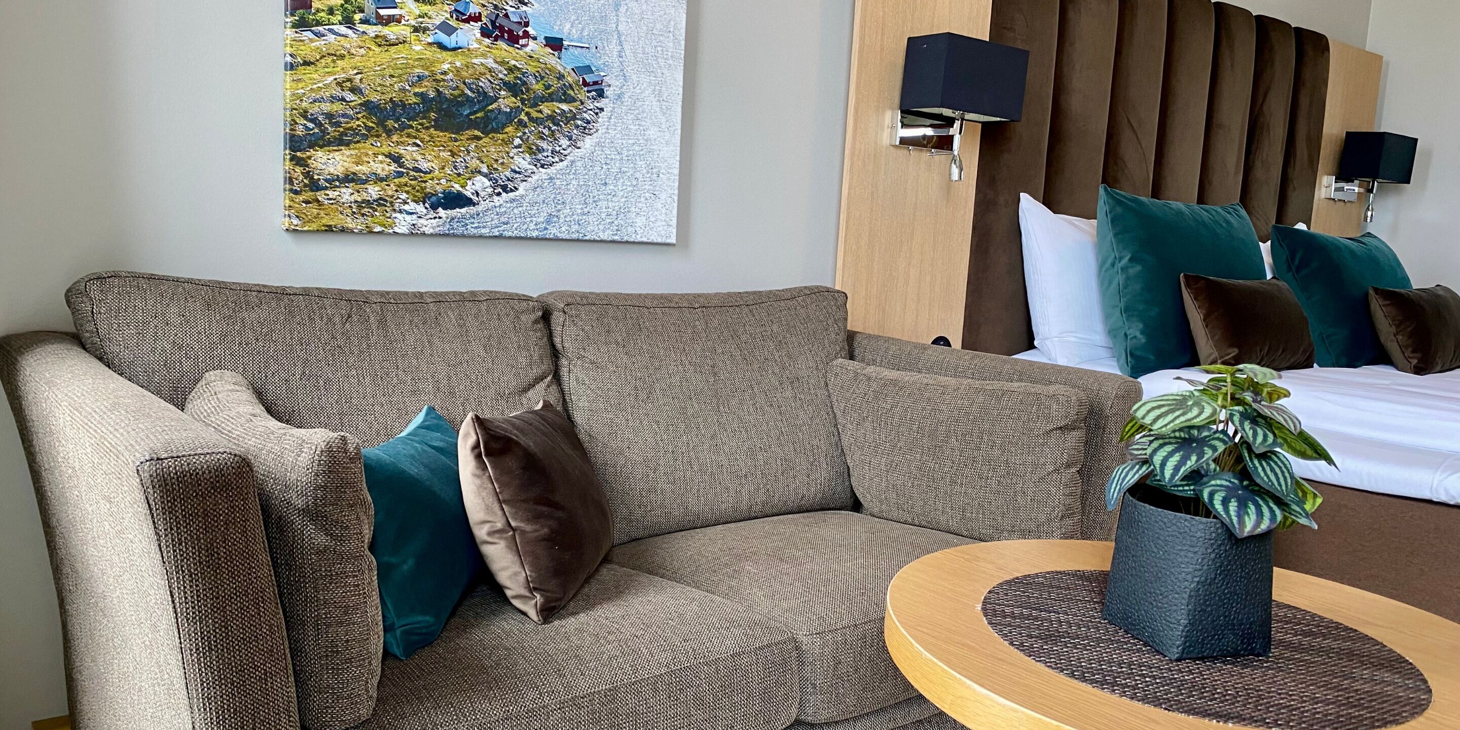 Habitación de hotel con un sofá marrón, almohadas verde azulado y marrón, una mesa de centro redonda de madera con una planta, un cuadro de un pueblo costero y una cama con almohadas verde azulado y marrón.