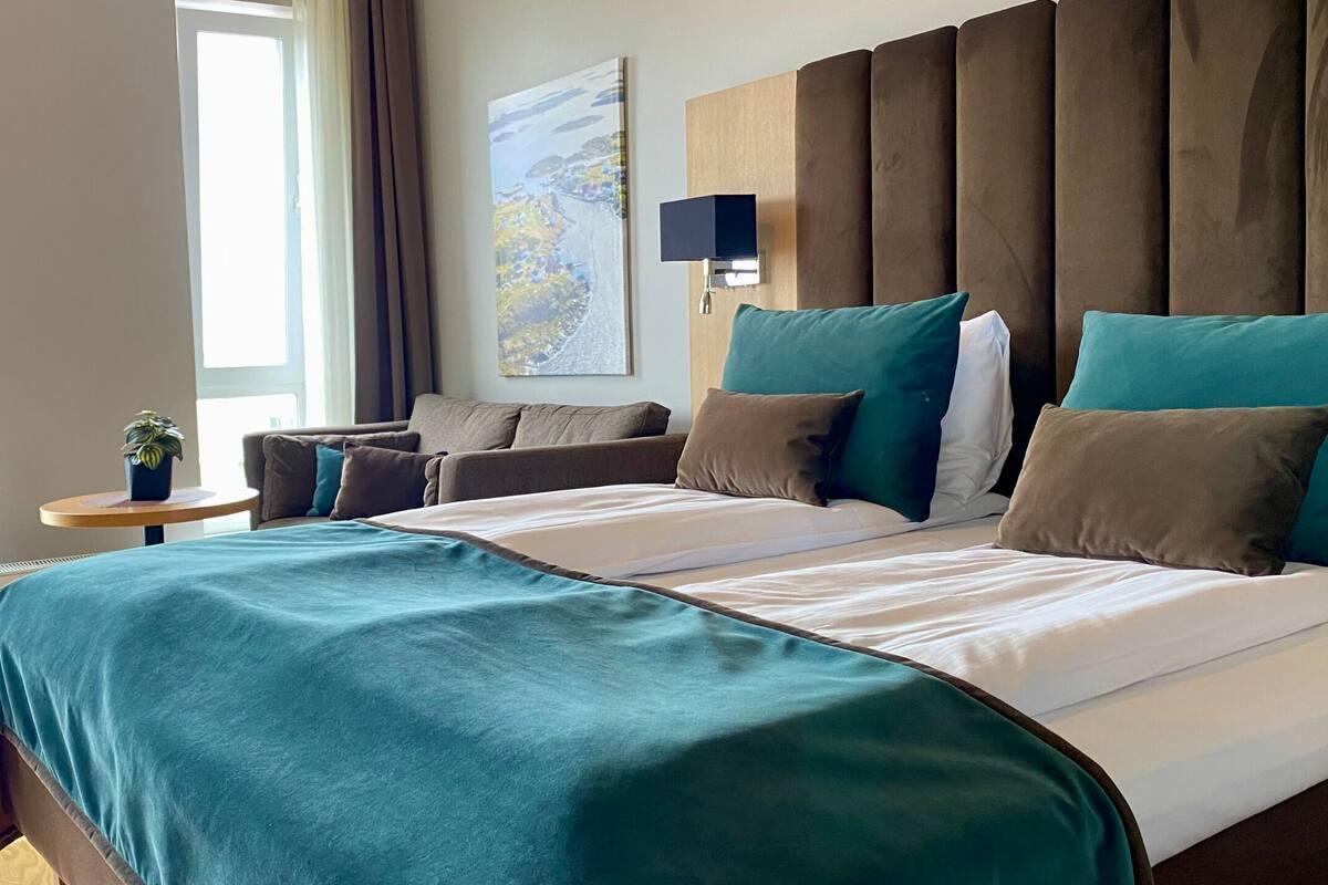 A modern hotel room with a large bed, a sofa, a small table with a plant, and a window with a view.