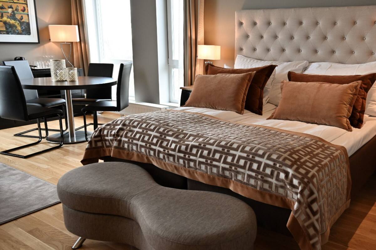 A modern hotel room with a large bed, a sitting area with a table and chairs, and a small desk.