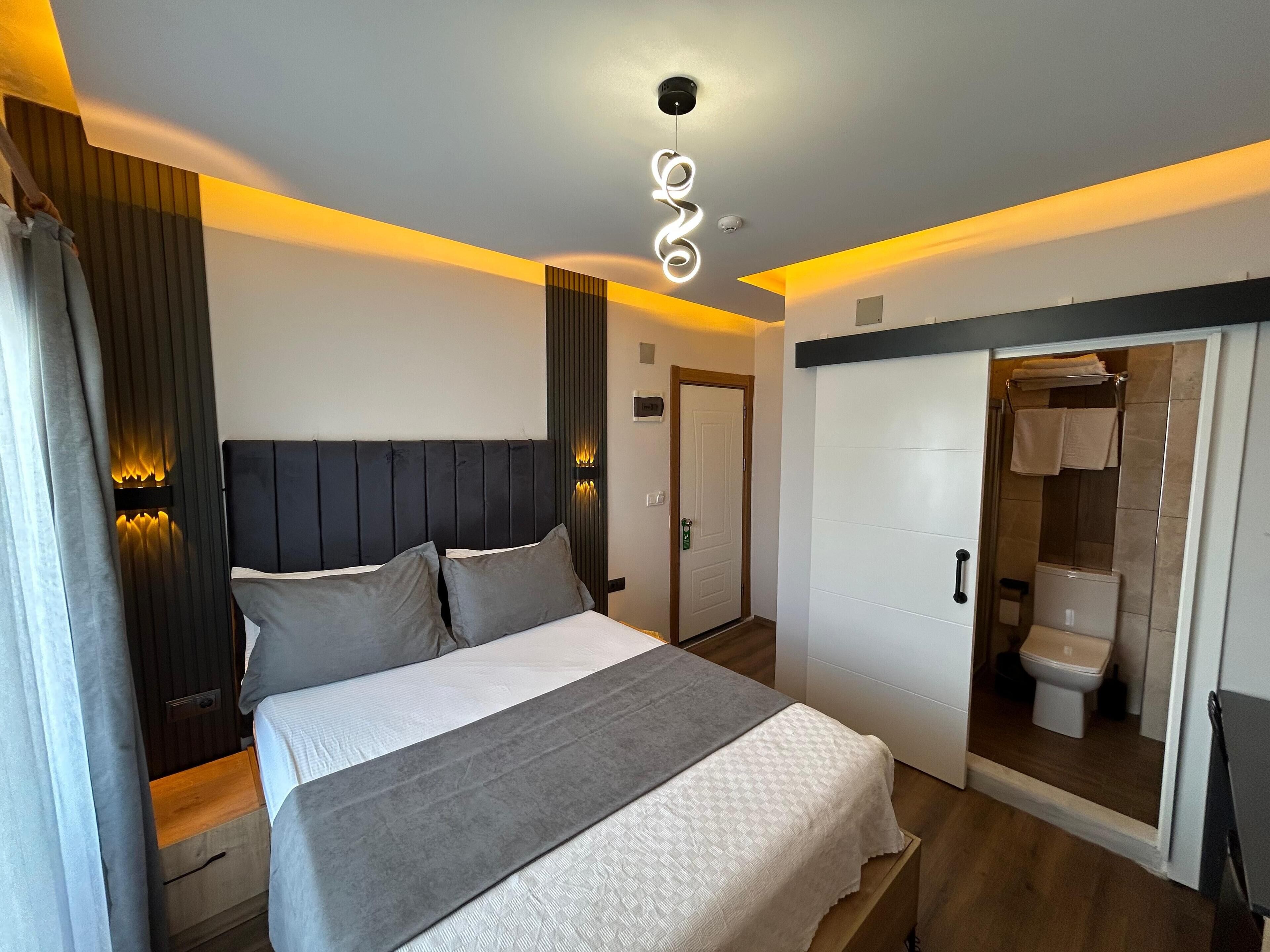 Un dormitorio moderno con una cama grande, mesitas de noche, un armario y un baño visible a través de una puerta abierta.