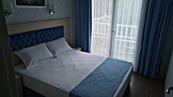 A neatly made bed with a blue headboard, a bedside table with a lamp, and a window with blue curtains.