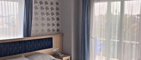 A hotel room with a large bed, a wooden headboard, a nightstand, a telephone, and a view of the outside through sheer curtains.